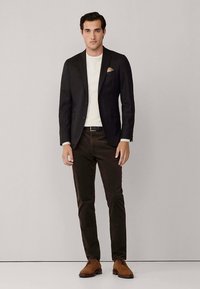 Zwarte blazer, wit overhemd, bruine broek en bruine schoenen. Gestructureerde stof met detail van een pochet. Model staat tegen een neutrale achtergrond.