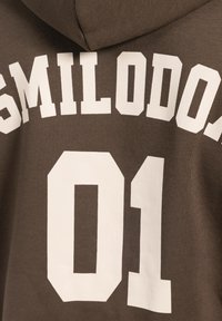 Brauner Hoodie mit großem weißen Text, der "SMILODO" über "01" liest, aus einem weichen, strukturierten Stoff. Die Kapuzenakzente sind sichtbar.