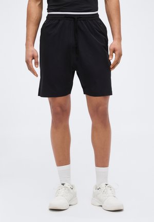 Homme portant un short noir à cordon, des chaussettes blanches montantes et des baskets blanches, debout devant un fond clair uni.