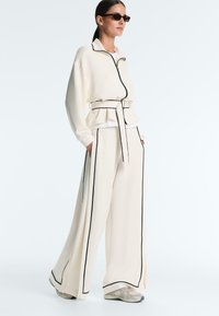 Femeie purtând pantaloni largi de culoare crem și jachetă cu fermoar cu bordură neagră, curea, adidași și ochelari de soare dreptunghiulari, stând din profil.