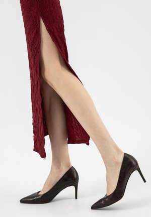 Vrouw met zwarte, gestructureerde hoge hakken en een lange bordeauxrode jurk met een zijsplitsing, waarbij de benen zichtbaar zijn tegen een effen achtergrond.
