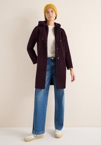 Bordeaux kleurige jas met een capuchon, voorzien van een zachte voering, twee knopen aan de voorkant, en brede mouwen, gecombineerd met lichtblauwe jeans en beige sneakers.