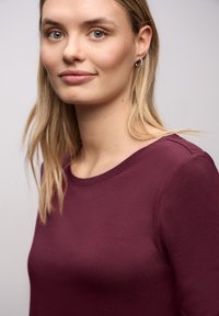 Bordowa bluzka z okrągłym dekoltem, wykonana z gładkiej tkaniny, z długimi rękawami i dopasowanym krojem. Styl minimalistyczny, bez wzorów i ozdób.