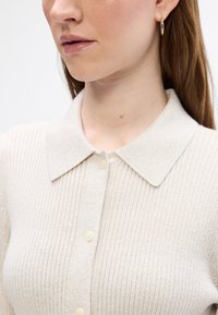Lys beige ribbet cardigan med en strukturert krage, knappelukking og myk tekstur. Har en subtil glans og en formsydd silhuett.