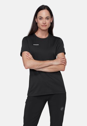 Kobieta z długimi ciemnymi włosami, ubrana w czarną koszulkę i spodnie Mammut, stojąca z założonymi na siebie ramionami na jednolitym tle.