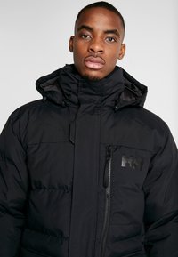 Black puffer jacket with a high collar, zip closure, and a chest pocket. Features a matte logo and a smooth, textured fabric finish.