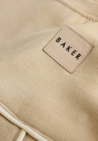 Beżowa tkanina o fakturowanej powierzchni posiada wyszywaną kwadratową metkę z napisem "BAKER" w czerni. Metka ma kontrastującą krawędź.