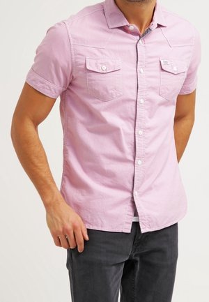 Man draagt een lichtroze kortärmelig blouse met knopen en twee borstzakken, gecombineerd met donkergrijze broek, tegen een effen achtergrond.