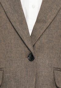 Hnedý textúrovaný blazer zapnutý nad bielou košeľou s golierom, ktorý zobrazuje klopy a horné vrecká.