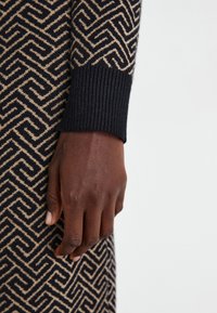 Suéter de malha com padrão geométrico em azul escuro e bege. Close-up da manga canelada e mão contra o tecido com padrão.