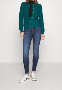 Kvinna i teal-färgad knappad cardigan, svart polotröja, blå skinny jeans och vita låga Converse-sneakers stående mot en enfärgad bakgrund.