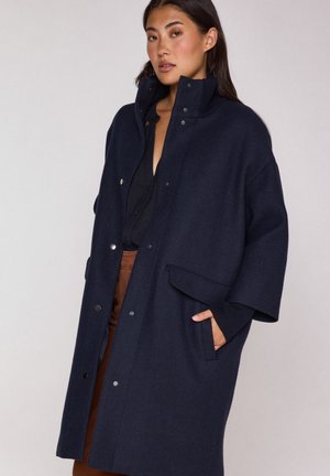 Femme portant un manteau oversized bleu marine foncé avec des boutons-pression et un col haut, posant avec une main dans la poche sur un fond clair uni.