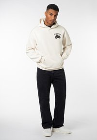 Beige hooded sweatshirt with front pocket, featuring a motorcycle graphic and text on the left chest, paired with black jeans and white sneakers.