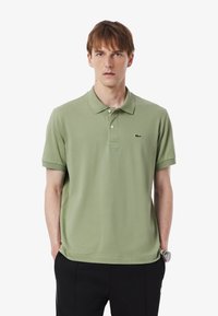 Polo shirt verde in cotone con maniche corte, colletto classico, chiusura a tre bottoni e logo ricamato sul lato sinistro del petto. Indossato con pantaloni neri.