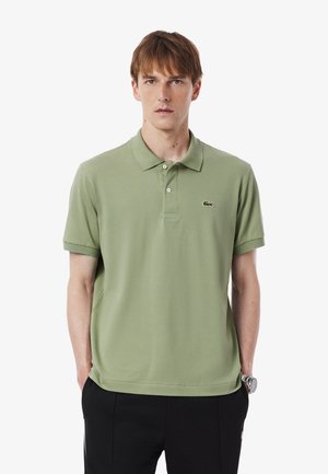 Polo shirt verde in cotone con maniche corte, colletto classico, chiusura a tre bottoni e logo ricamato sul lato sinistro del petto. Indossato con pantaloni neri.