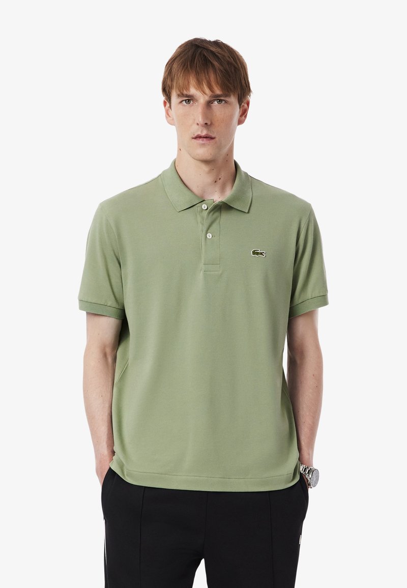Green cotton polo shirt with short sleeves, classic collar, three-button placket, and embroidered logo on the left chest. Worn with black pants.