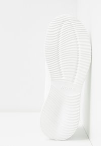 Vit skosula med texturerade spår och varumärket "BOBS" präglat, lutande mot ett hörn. 