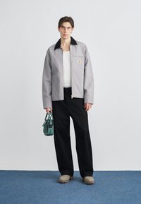 Jachetă gri cu guler negru, fermoar frontal și buzunar la piept. Asortată cu pantaloni negri cu croială largă și clape bej. Ține o geantă mică verde.