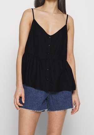 Frau trägt ein schwarzes, ärmelloses Top mit Knöpfen und dünnen Trägern sowie blaue Jeans-Shorts mit ausgefranstem Saum.