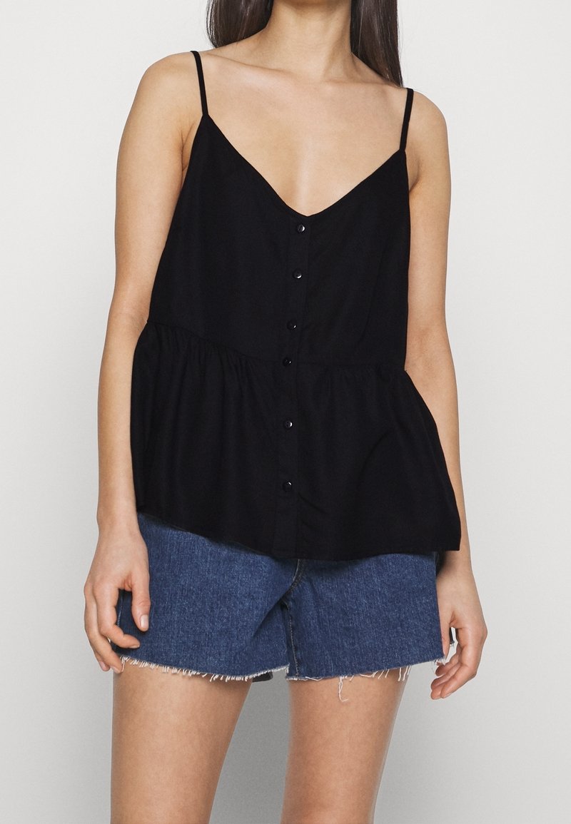 Femme portant un haut noir sans manches boutonné avec des fines bretelles et un short en jean bleu aux bords effilochés.