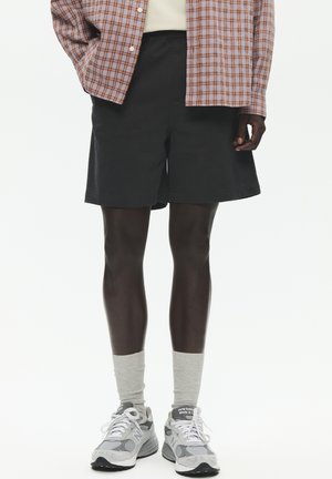 Person iført sorte shorts, grå sneakers med hvide snørebånd, grå mellemlange sokker og en brunternet skjorte over en hvid top.