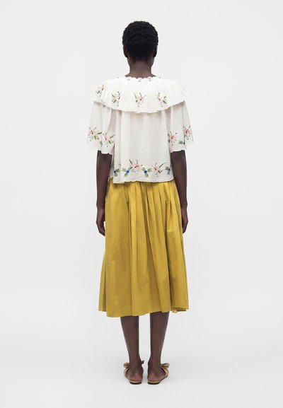 Femme debout de dos, portant un chemisier blanc brodé de fleurs avec un col à volants et une jupe midi plissée jaune avec des sandales.