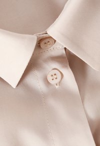 Beige button-up shirt with a smooth texture, featuring a classic collar and two light buttons near the neckline.