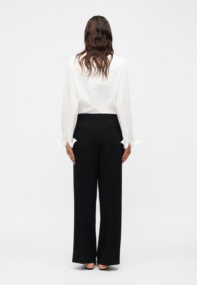 Γυναίκα με μακριά κυματιστά μαλλιά, φοράει μια λευκή μπλούζα με μακριά μανίκια και μαύρο παντελόνι με φαρδιά πόδια, στέκεται γυρισμένη πλάτη σε απλό λευκό φόντο.