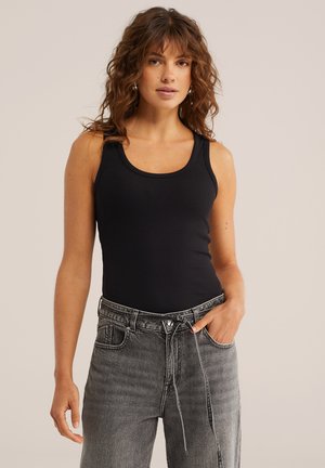 Kobieta o falowanych brązowych włosach, ubrana w czarny, bezrękawnik i szare, wysokie spodnie dżinsowe, z lewą ręką w kieszeni, na jednolitym tle.