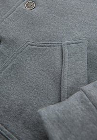 Šedá mikina s hladkým, texturovaným materiálem. Disponuje zapínáním na knoflíky vpředu, velkou kapsou a žebrovanými manžetami.