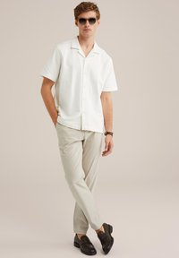 Camicia bianca a maniche corte con texture e colletto, pantaloni beige chiaro e mocassini scuri. Modello che indossa occhiali da sole, in piedi con una mano in tasca.