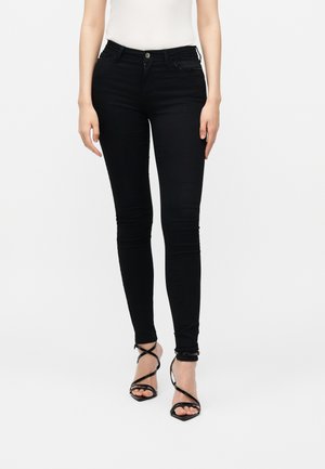 Kvinde iført sorte skinny jeans og sorte højhælede sandaler med stropper, stående mod en ensfarvet hvid baggrund.
