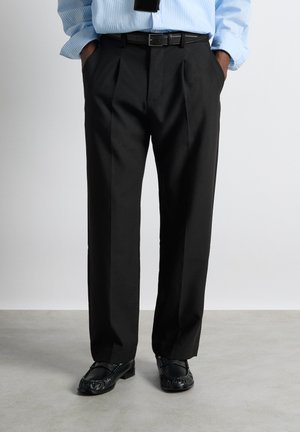 Mann trägt schwarze plissierte Anzughose, schwarzen Gürtel, schwarze Slipper und hellblau gestreiftes Langarmhemd mit Händen in den Taschen.