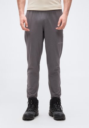 Muž oblečený v sivých vonkajších nohaviciach Columbia a čiernych turistických topánkach, stojaci s rukami uvoľnene pozdĺž tela na bielom pozadí.