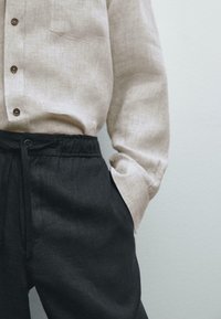 Light beige linen shirt with a chest pocket, buttoned up to the neck, paired with dark gray linen drawstring pants.