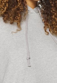 Haut à capuche gris en tissu doux. Il possède une capuche avec cordon de serrage et une surface texturée. La couleur est uniforme, sans motifs ni embellissements visibles.