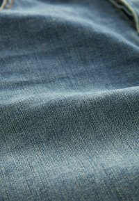 Tejido de mezclilla, azul medio con una textura sutil, que muestra un clásico tejido de sarga. Superficie suave y lisa, con un ligero desgastado y sin patrones.