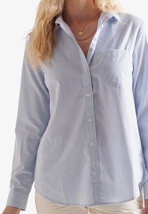 Kobieta ubrana w błękitno-białą koszulę w cienkie prążki z kieszonką na piersi oraz warstwowe złote naszyjniki.