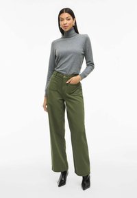 Maglione grigio a collo alto, pantaloni larghi verde oliva con tasche frontali e stivali neri a punta alla caviglia. Design semplice e pulito, con una texture liscia.