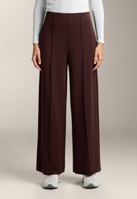 Bordowe spodnie o szerokich nogawkach, wykonane z miękkiego materiału, z plisami, stylizowane z białą bluzką z długim rękawem i szarymi sneakersami.