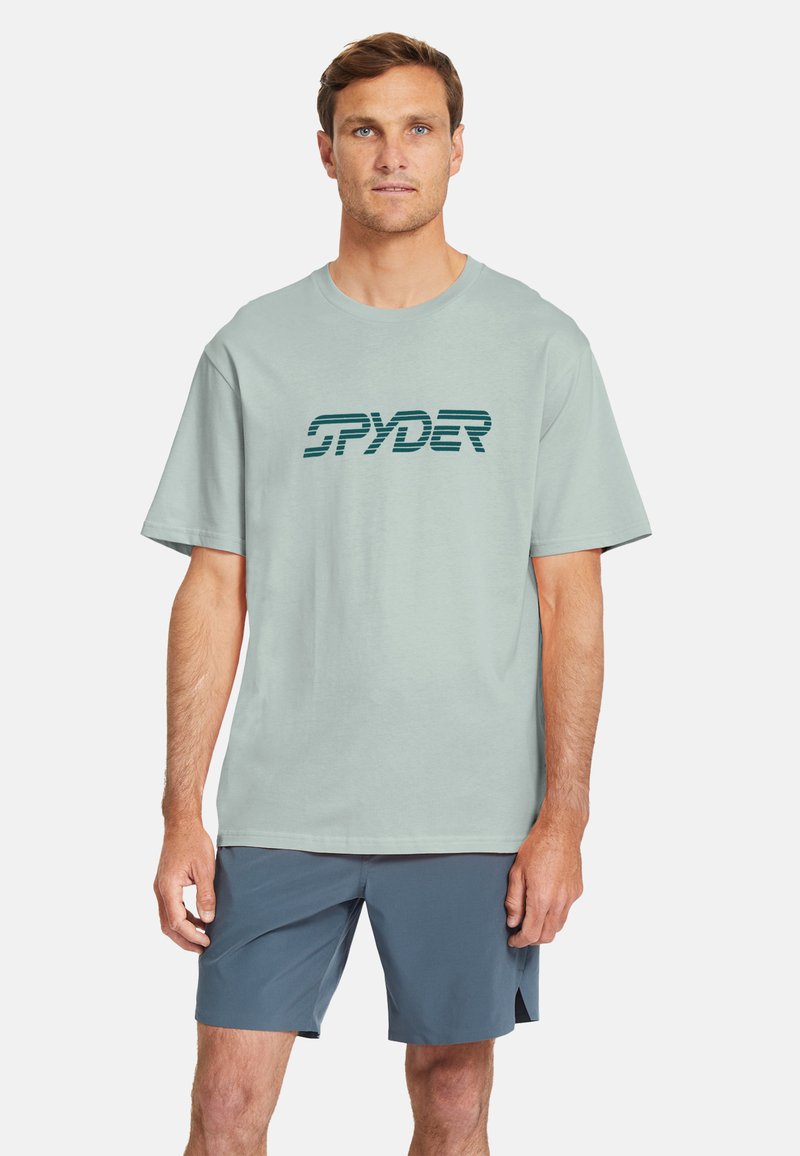 Camiseta de algodón verde claro con el logo "SPYDER" en verde oscuro, corte relajado, de mangas cortas. Combinada con pantalones cortos azul oscuro.