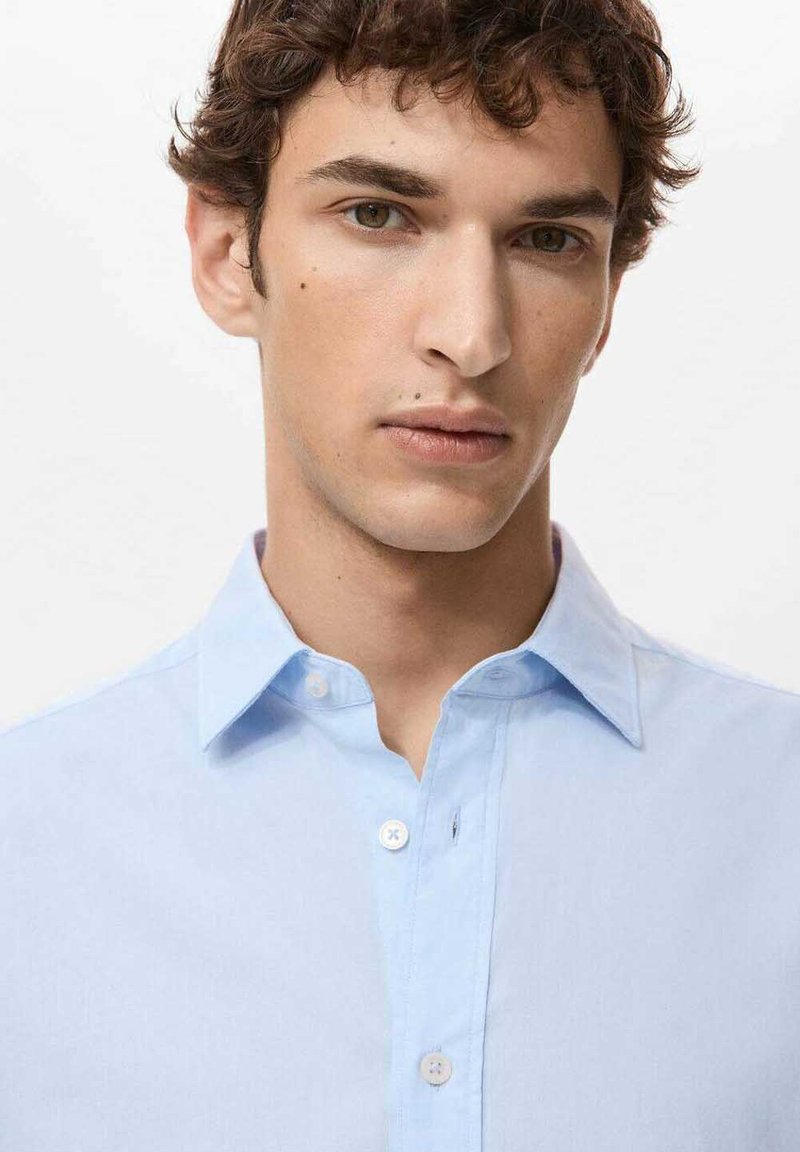 Camicia azzurro chiaro con bottoni in tessuto liscio, colletto standard, caratterizzata da una pattina con bottoni bianchi e design minimalista.