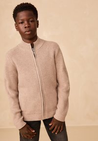 Pull beige clair zippé avec texture côtelée et col montant, en tissu épais tricoté avec des poignets ajustés, associé à un jean gris foncé.