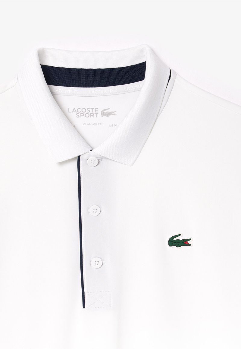 Chemise polo blanche avec un col marine, trois boutons et un logo de crocodile vert. Présente un tissu texturé pour un look sportif.