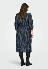 Vestido azul marino con un diseño fluido, que presenta un patrón ondulado en dorado, cintura elástica y mangas tres cuartos. Lleva consigo botas negras hasta el tobillo.