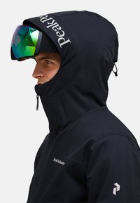 Schwarze wasserdichte Skijacke mit hohem Kragen, verstellbarer Kapuze und Peak Performance Logo. Glatte Textur mit einem Reißverschluss vorne.