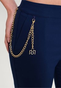 Detailní záběr na zlatý řetěz připevněný k kapse námořnických modrých kalhot, s klipem a ozdobným přívěskem "RR".