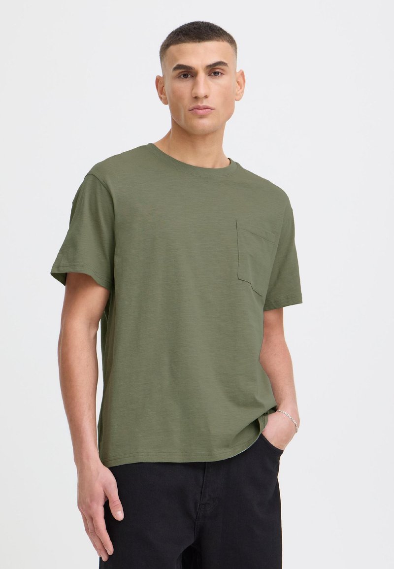 Hombre con pelo corto que lleva una camiseta de manga corta verde oliva con un bolsillo en el pecho y pantalones negros sobre un fondo claro y liso.