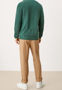 Homme portant un pull en tricot vert, un pantalon brun clair retroussé, des chaussettes blanches et des baskets blanches, debout de dos sur un fond neutre.