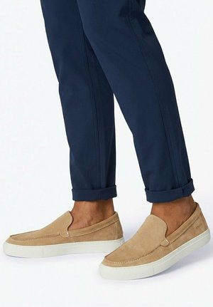 Béžové semišové topánky bez šnúrok s bielymi podrážkami nosené s tmavomodrými nohavicami s manžetami na svetlom podklade.